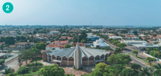 Imagem: Fotografia. Uma igreja no meio de uma praça com árvores. Ao redor, a cidade. Fim da imagem.