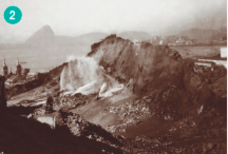 Imagem: Fotografia. Um morro com partes destruídas. Ao lado tem a torre de uma igreja.  Fim da imagem.