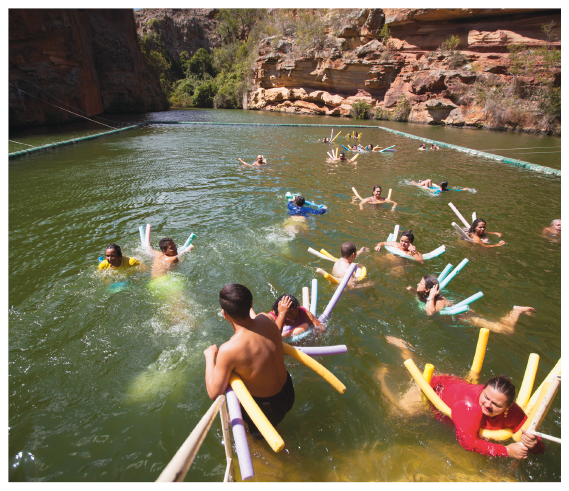 Imagem: Fotografia. Pessoas nadando em um rio. Elas estão apoiadas em cilindros de isopor boiando.  Fim da imagem.