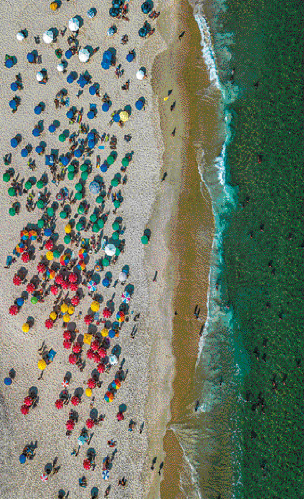 Imagem: Fotografia. Uma praia vista de cima. Na parte da areia tem pessoas caminhando e muitos guarda-sóis coloridos. A água está na direita e há pessoas nadando.  Fim da imagem.