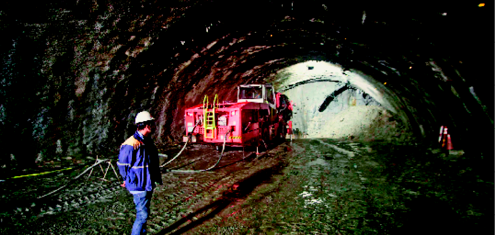 Imagem: Fotografia. Homem de jaqueta azul e capacete de proteção parado com as mãos para trás em um túnel de barro. Ao lado dele, um caminhão vermelho.  Fim da imagem.