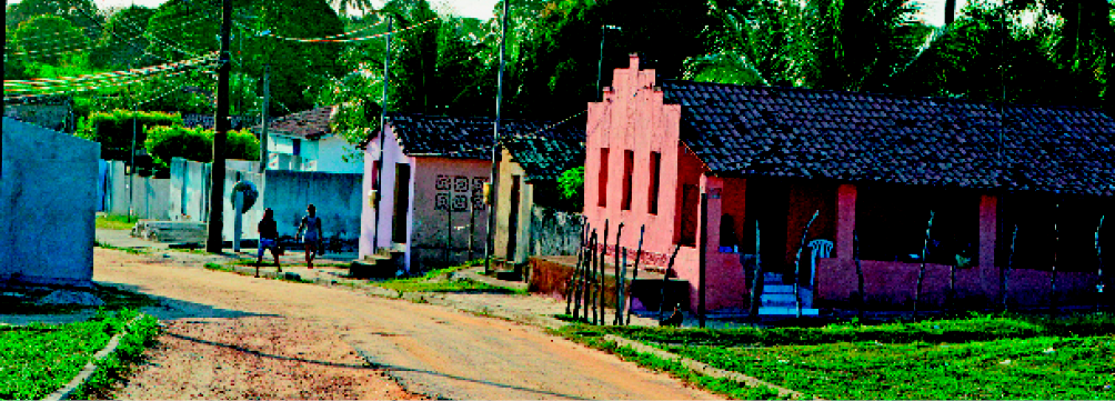 Imagem: Fotografia. Uma rua de terra com casas e um lote gramado de um dos lados. Na calçada há duas mulheres.  Fim da imagem.