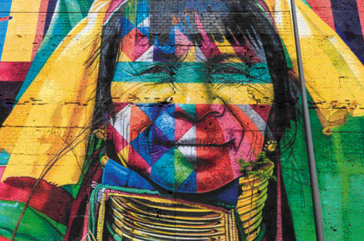 Imagem: Fotografia. Muro com pintura colorida do rosto de mulheres indígenas. O rosto está pintado com faixas coloridas: verde, vermelho, amarelo, azul e laranja. A mulher olha para a frente e sorri.  Fim da imagem.