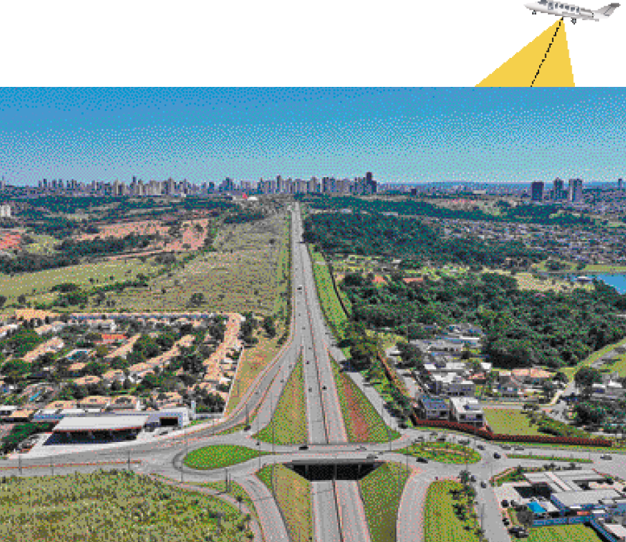 Imagem: Fotografia. Cruzamento de duas avenidas, com duas rotatórias. Ao redor, áreas de construção e áreas de vegetação. Acima, um avião voando inclinado com uma linha na diagonal até a fotogra-fia.  Fim da imagem.