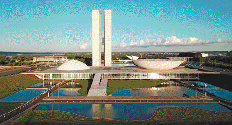 Imagem: Fotografia. Vista aérea de duas torres altas e de cada lado uma construção com o formato de uma parte de uma esfera. Na frente, um lago e áreas gramadas.  Fim da imagem.