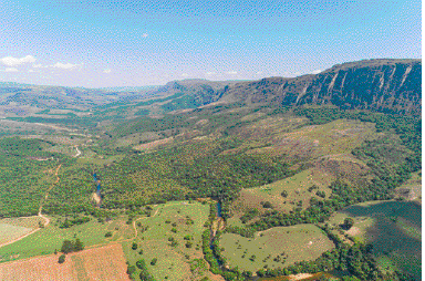 Imagem: Fotografia. Paisagem com campos com vegetação e montanhas baixas no fundo.   Fim da imagem.