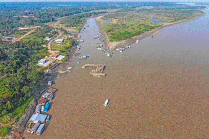 Imagem: Fotografia. Um rio extenso com a água marrom. Nas margens tem barcos e dos lados áreas com árvores.   Fim da imagem.