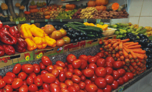 Imagem: Uma banca com caixas de papelão com diversas frutas e legumes: tomates, pimentões, cenouras e pepino.   Fim da imagem.