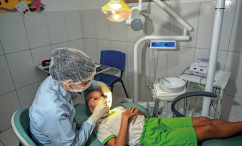 Imagem: Fotografia. Um menino de bermuda verde e camiseta branca sentado em uma cadeira de dentis-ta. Ao lado dele, uma mulher de touca, luva e jaleco olha para a boca do menino.   Fim da imagem.