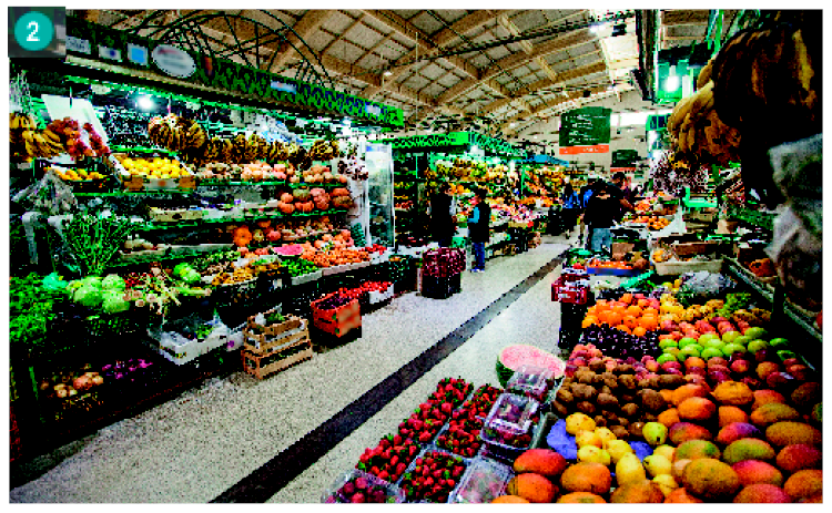 Imagem: Fotografia. Um corredor com barracas de frutas e legumes dos dois lados. As frutas e legumes estão separados em caixas.  Fim da imagem.