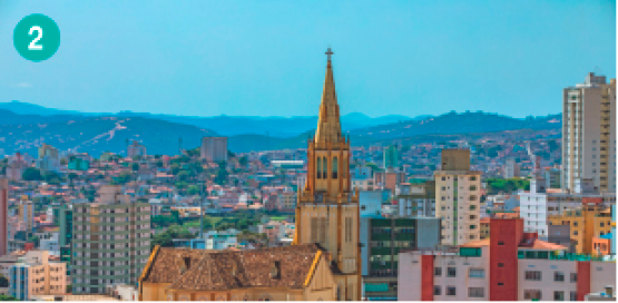 Imagem: Fotografia. Vista aérea de uma cidade com prédios e uma torre de igreja amarela.  Fim da imagem.