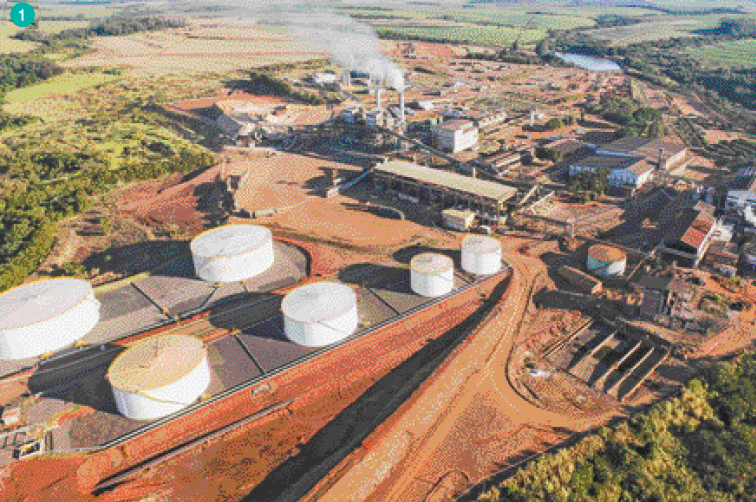 Imagem: Fotografia. Uma fábrica no meio de uma área rural. Na parte da esquerda há seis tanques cilíndri-cos. Na parte da direita há construções com chaminés saindo fumaça.  Fim da imagem.