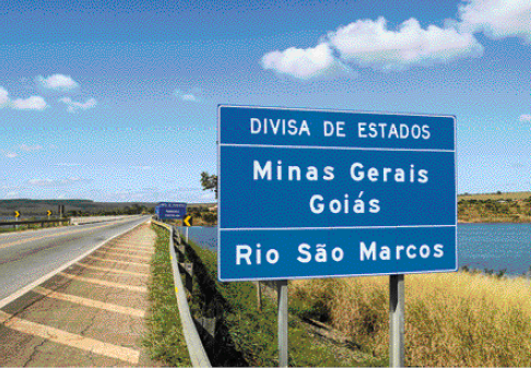 Imagem: Fotografia. Uma placa azul retangular na beira da estrada. Texto na placa: divisa de estados. Mi-nas Gerais, Goiás e Rio São Marcos.   Fim da imagem.