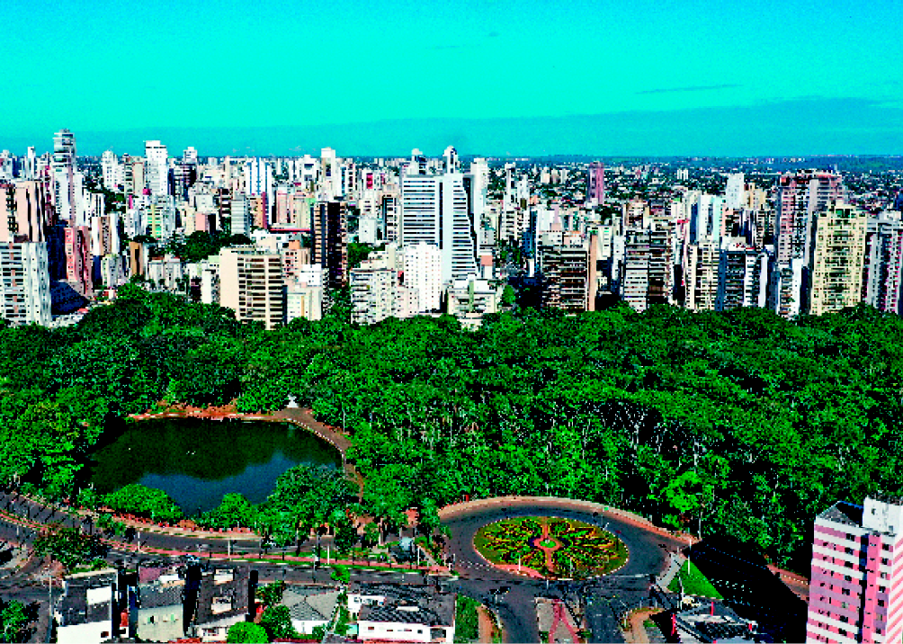 Imagem: Fotografia. Vista aérea de uma paisagem de parque com árvores e um lago. Na frente, uma rota-tória e uma avenida. Ao fundo, prédios.  Fim da imagem.