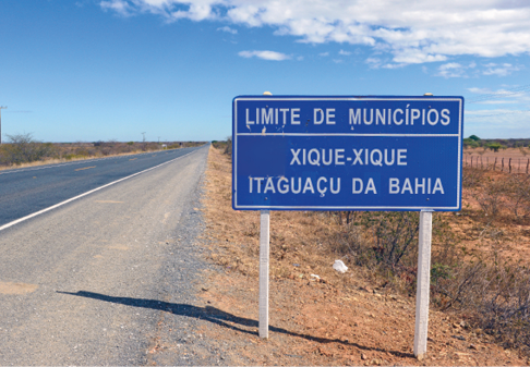 Imagem: Fotografia. Uma placa azul retangular na beira da estrada. Texto na placa: limite de municípios. Xique-xique e Itaguaçu da Bahia.   Fim da imagem.