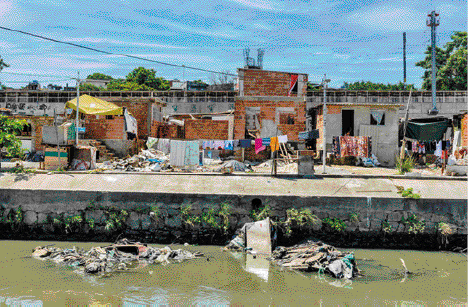 Imagem: Fotografia. Uma comunidade com casas de tijolos construídas na margem de um rio. O rio está poluído, com pilhas de lixo. Na frente das casas tem varais com roupas penduradas.  Fim da imagem.