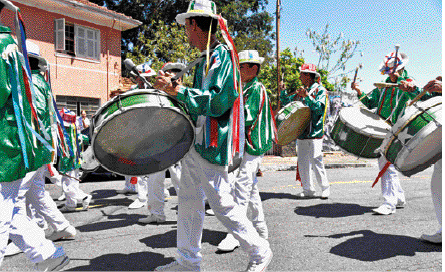 Imagem: Fotografia. Um grupo de homens com calça branca, camisa verde listrada e chapéu branco com fitas. Eles estão em uma rua e tocam tambor.   Fim da imagem.