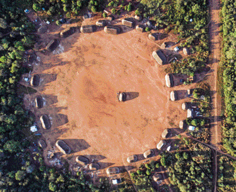 Imagem: Fotografia. Vista aérea de uma vila com ocas. Elas estão enfileiradas lado a lado formando um grande círculo ao redor de um terreno de terra. No centro desse terreno tem outra oca. Ao re-dor, área coberta por árvores.  Fim da imagem.