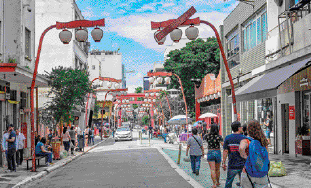 Imagem: Fotografia. Uma rua com pessoas caminhando nas calçadas. Nas calçadas há postes vermelhos de iluminação com três lâmpadas em cada. Fim da imagem.