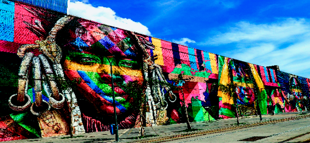 Imagem: Fotografia. Um grande muro com pintura colorida de três rostos de mulheres indígenas. Os ros-tos estão pintados com faixas coloridas: verde, vermelho, amarelo, azul e laranja. Na calçada na frente do muro há um poste de iluminação com uma lixeira e árvores.  Fim da imagem.