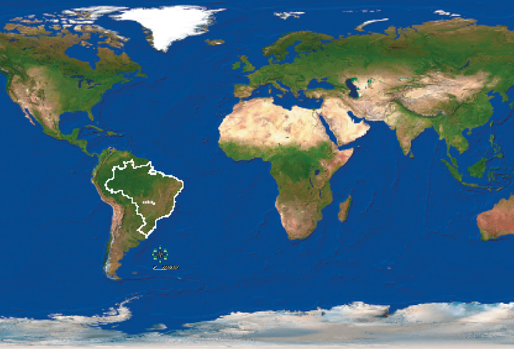 Imagem: Fotografia. Uma menina de cabelo castanho liso e longo. Ela está sorrindo e sentada de pernas cruzadas. Ela usa uma calça cinza e uma camiseta branca. Atrás dela, imagem do mapa mundi, com os continentes em verde e marrom e o oceano em azul. O Brasil está destacado.  Fim da imagem.