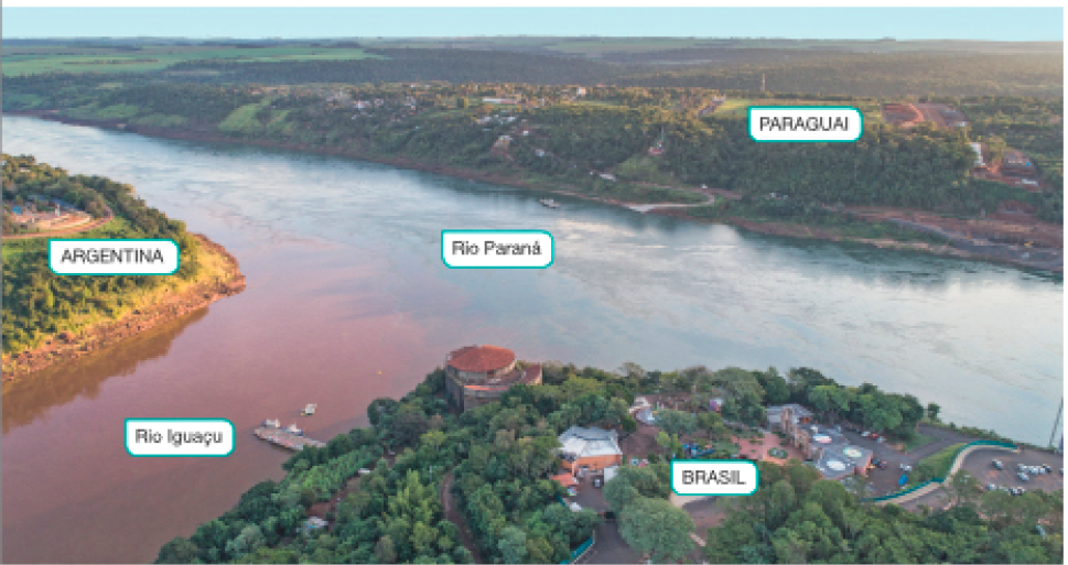 Imagem: Fotografia. Paisagem mostrando o encontro de dois rios: Rio Iguaçu e Rio Paraná. Nas margens dos rios, forma-se três blocos de terra, cada um é um país: Paraguai, Argentina e Brasil.   Fim da imagem.
