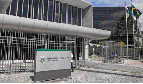 Imagem: Fotografia. Fachada da Assembleia Legislativa do Paraná. Um prédio cinza com janelas grandes de vidro e uma grade branca na frente.   Fim da imagem.