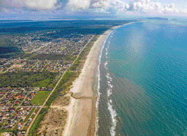 Imagem: Fotografia. Uma praia com o mar na direita e uma cidade na esquerda.   Fim da imagem.