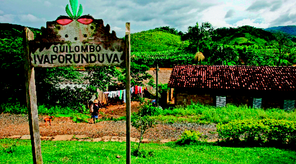 Imagem: Fotografia. Uma paca de madeira com o desenho de olhos e o texto: Quilombo Ivaporunduva. No fundo, uma casa de tijolos com um varal de roupas na frente e no fundo montanhas.  Fim da imagem.