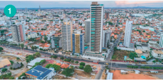 Imagem: Fotografia. Vista aérea de uma cidade com uma avenida em primeiro plano e alguns prédios es-paçados.  Fim da imagem.