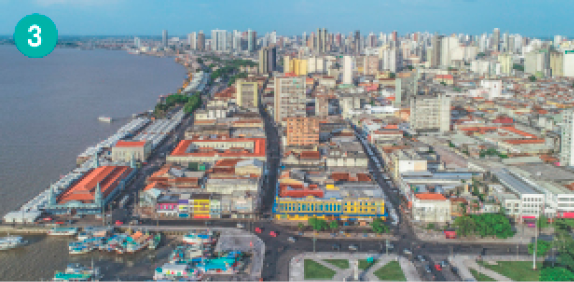 Imagem: Fotografia. Vista aérea de uma cidade com casas na parte da frente e prédios no fundo., A cidade está ao lado do mar.  Fim da imagem.