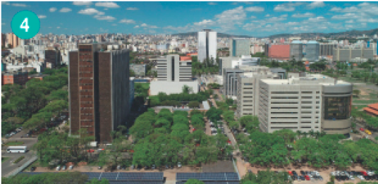 Imagem: Fotografia. Vista aérea de uma cidade com prédios e grandes áreas com árvores.  Fim da imagem.