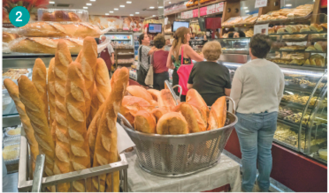 Imagem: Fotografia. Em primeiro plano, duas cestas de metal com pães. Atrás, uma fila de pessoas em uma padaria. Ao lado delas há um balcão com pães.   Fim da imagem.