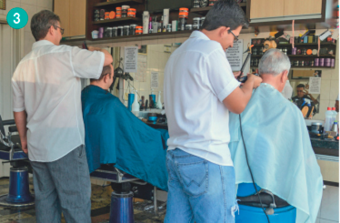 Imagem: Fotografia. Uma barbearia com dois homens sentados em cadeiras e dois cabelereiros, um atrás de cada uma.  Fim da imagem.