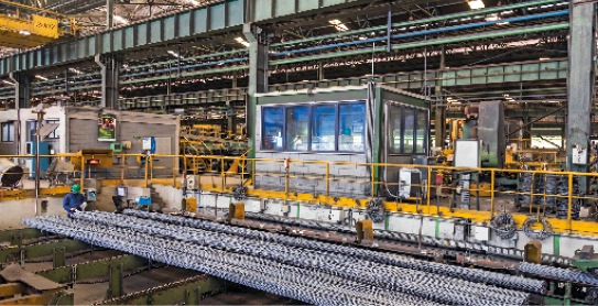 Imagem: Fotografia. Interior de uma fábrica, com grandes estruturas metálicas e máquinas. Na parte da frente, barras de aço. Fim da imagem.