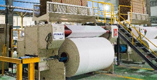 Imagem: Fotografia. Interior de uma fábrica com uma grande máquina com um rolo de papel.  Fim da imagem.