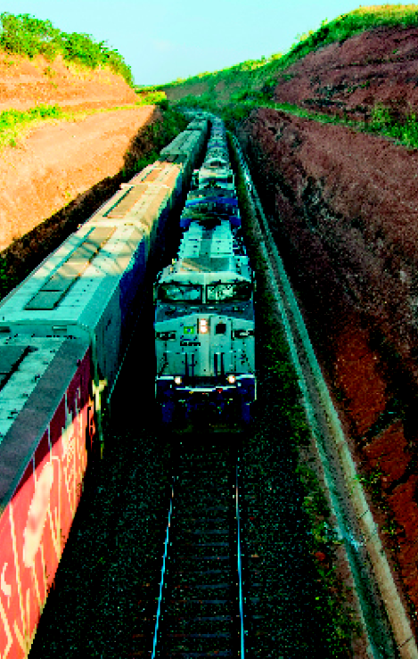 Imagem: Fotografia. Dois trens em linhas lado a lado. Eles estão no meio de taludes de terra.  Fim da imagem.