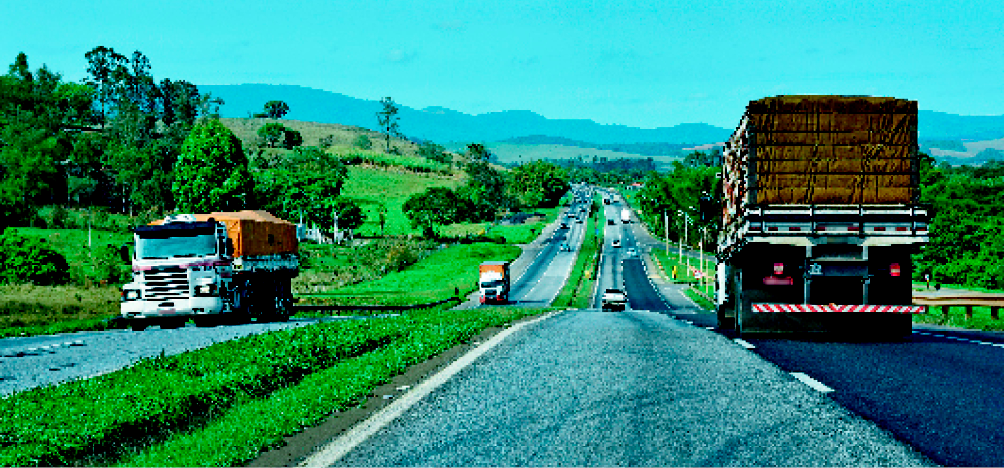Imagem: Fotografia. Uma estrada com caminhões indo nos dois sentidos. Nas margens da estrada: área coberta por vegetação.  Fim da imagem.