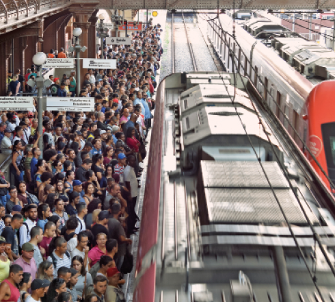 Imagem: Fotografia. Uma multidão em uma plataforma de trem. Há dois trens na linha.  Fim da imagem.
