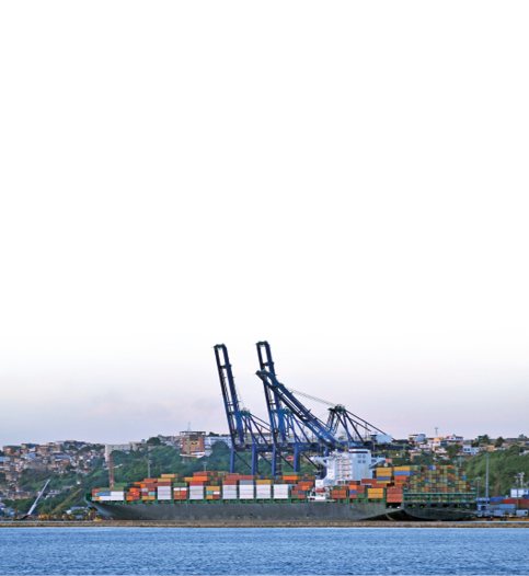 Imagem: Fotografia. Um navio atracado em um porto. Em cima, pilhas de containers retangulares de diferentes cores.  Fim da imagem.