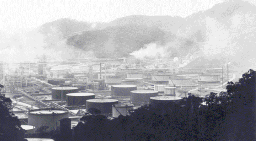 Imagem: Fotografia. Imagem em preto e branco de uma indústria, com tanques cilíndricos e no fundo chaminés com fumaça saindo. Atrás, montanhas.  Fim da imagem.