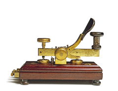 Imagem: Fotografia. Um telégrafo, máquina com a base de madeira escura e metal dourado em cima.  Fim da imagem.