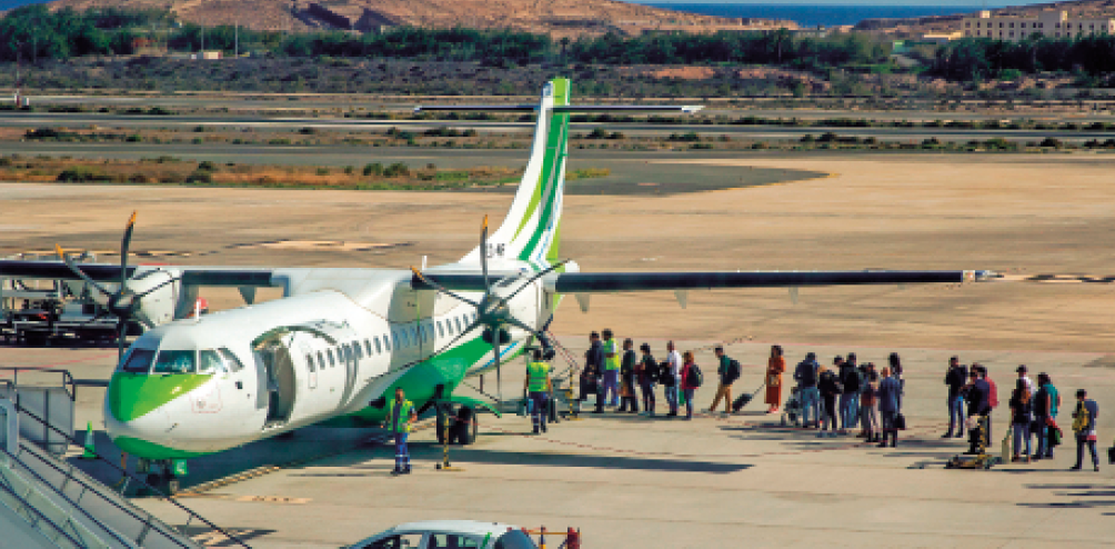 Imagem: Fotografia. Uma fila de pessoas na frente da porta de um avião branco e verde.  Fim da imagem.