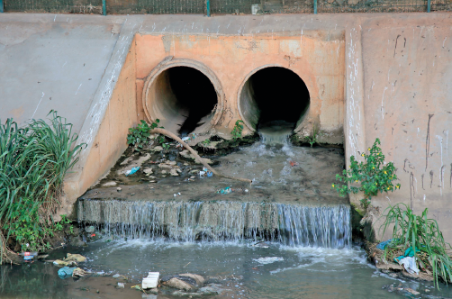 Imagem: Fotografia. Dois tubos de concreto com um rio na frente. Na ponta dos tubos há pilhas de lixo.  Fim da imagem.