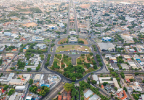 Imagem: Fotografia. Imagem aérea de uma cidade com uma grande praça no centro, com canteiros com árvores. As ruas da cidade saem da praça e as quadras tem casas.  Fim da imagem.