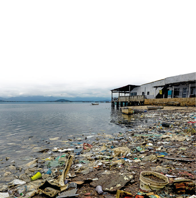 Imagem: Fotografia. Uma praia com lixo acumulado na água e na areia. Do lado, uma construção precária.  Fim da imagem.