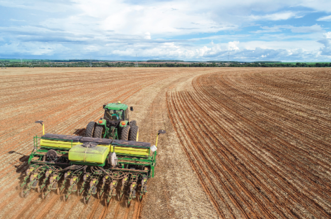 Imagem: Fotografia. Um trator verde passando em uma grande área de terra com plantação.  Fim da imagem.