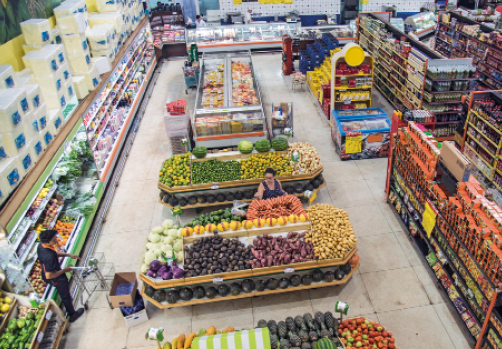 Imagem: Fotografia. Um supermercado visto de cima, com prateleiras com produtos diversos, geladeiras do lado esquerdo e ilhas com frutas no meio. Tem um homem de camiseta preta com um carrinho de supermercado e uma mulher de blusa regata pegando uma fruta.  Fim da imagem.