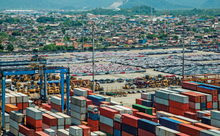 Imagem: Fotografia. Uma área com containers empilhados. Eles são de diversas cores e com tamanho padronizado. Ao fundo, uma cidade.  Fim da imagem.