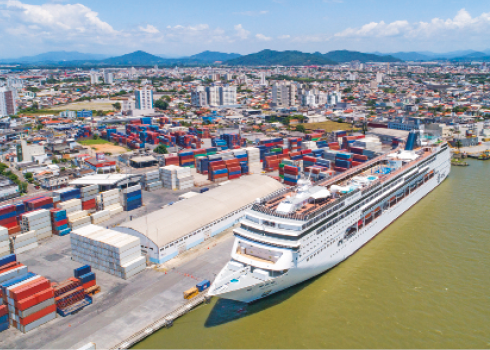 Imagem: Fotografia. Um navio atracado em um porto. Ao lado, um terreno com pilhas de containers retangulares.  Fim da imagem.